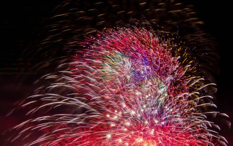 Bright, colorful fireworks burst against a dark night sky, creating vibrant streaks and patterns in red, white, blue, and green. The display fills the scene with light and energy.