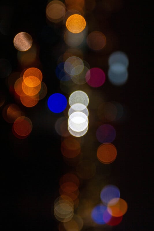 Out-of-focus multicolored lights, including white, orange, blue, pink, and yellow, creating circular bokeh against a dark background. The colors appear scattered and overlapping, forming an abstract pattern.