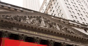 The facade of the New York Stock Exchange building, featuring classical columns and sculpted figures above the entrance, with tall skyscrapers in the background.