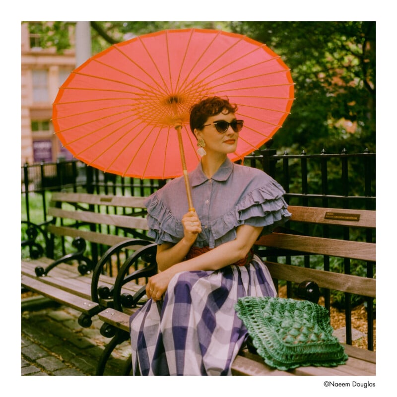 A woman in a ruffled blouse and checked skirt sits on a park bench, holding a large orange parasol. She wears sunglasses and has a green woven bag beside her. Trees and a black metal fence are in the background.