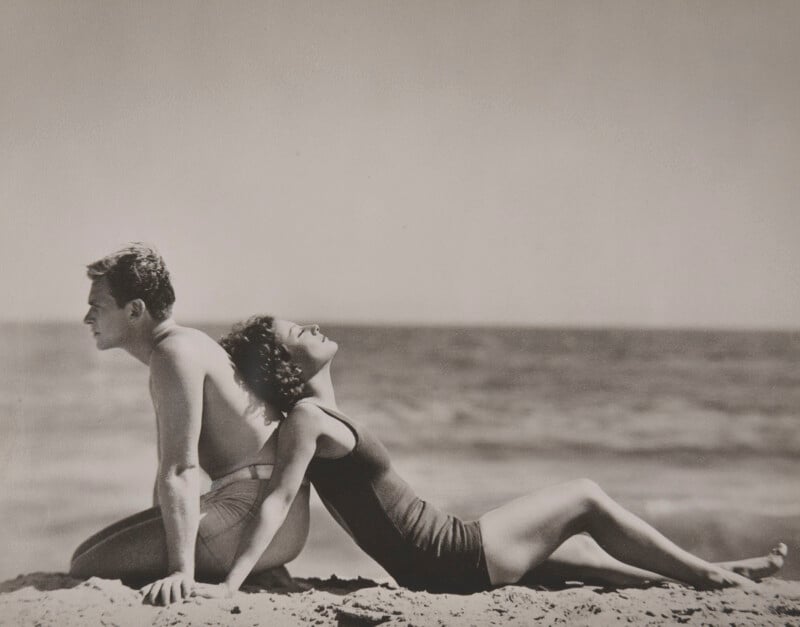 A man and woman in vintage swimsuits sit on the beach, leaning back to back, with the ocean in the background. The woman tilts her head back and smiles, enjoying the sun. The photo is in black and white.