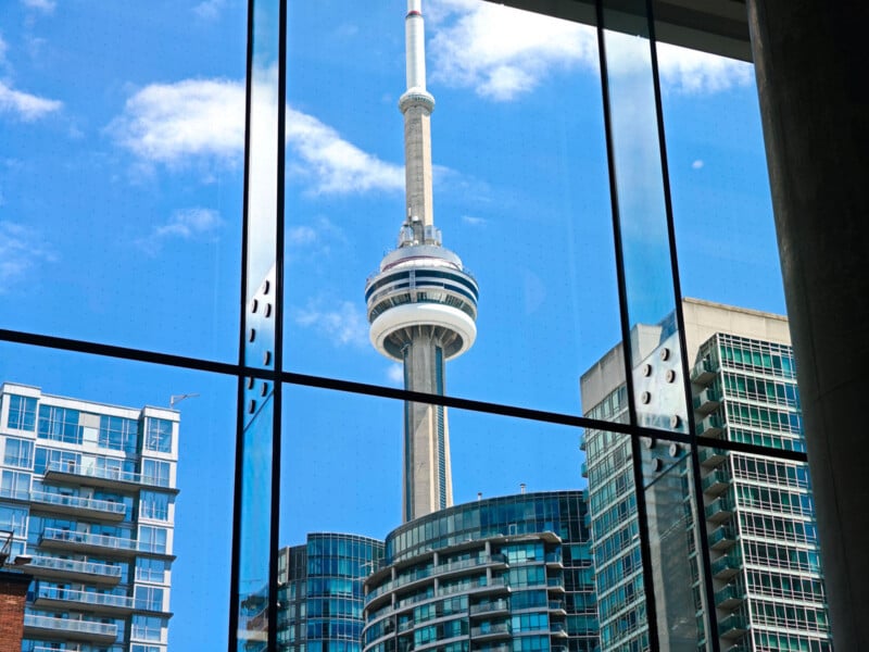 View of the CN Tower and surrounding modern buildings in Toronto, seen through large glass windows with reflections and blue sky in the background.