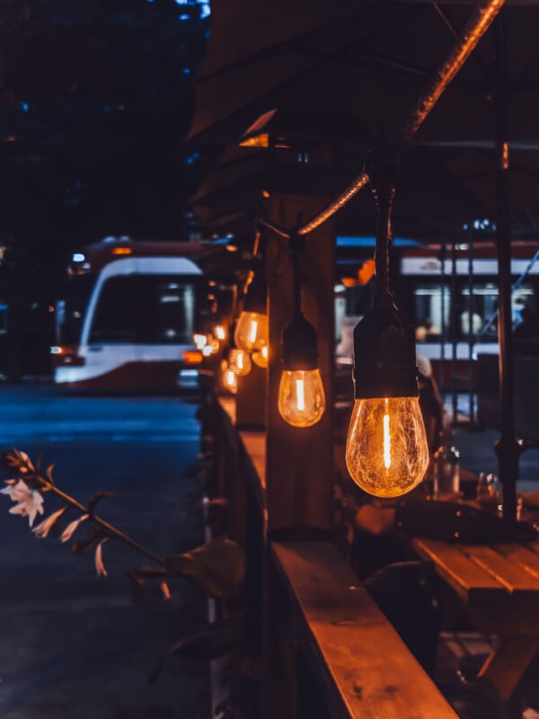 A row of glowing vintage-style light bulbs hangs along an outdoor patio at dusk, with blurred tables and chairs in the foreground and a passing streetcar in the background.