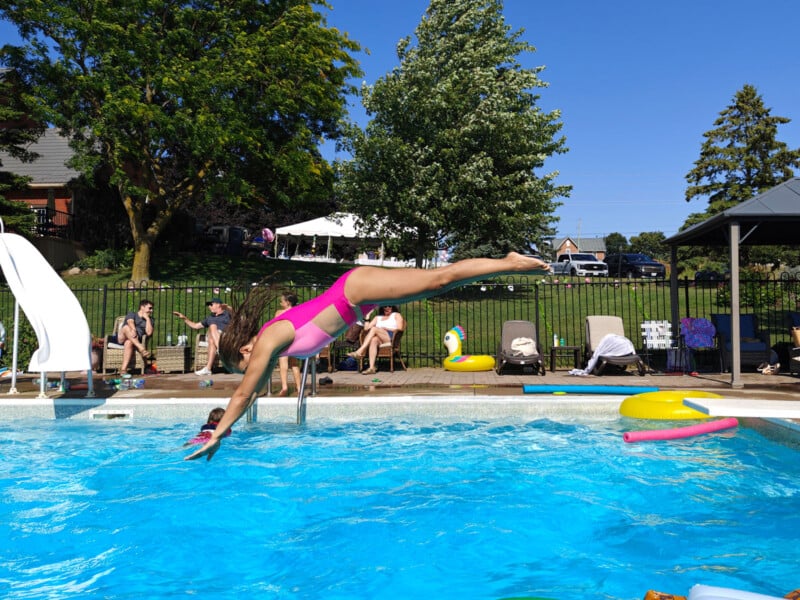 A person in a pink swimsuit dives into a bright blue swimming pool, with people relaxing and watching on lounge chairs and under a tent in the background, on a sunny day.