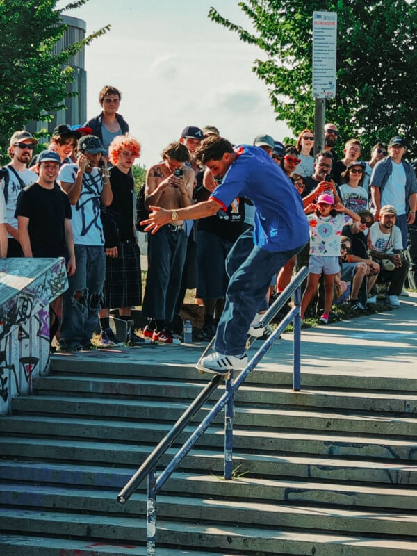 A skateboarder in a blue shirt grinds down a handrail at a skatepark, while a crowd of spectators watches and takes photos under sunny skies.