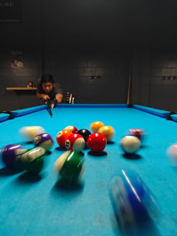 A person is taking a shot at a blue pool table, causing colorful billiard balls to scatter and blur from motion. The background is dimly lit, with gray walls and a shelf visible behind the player.