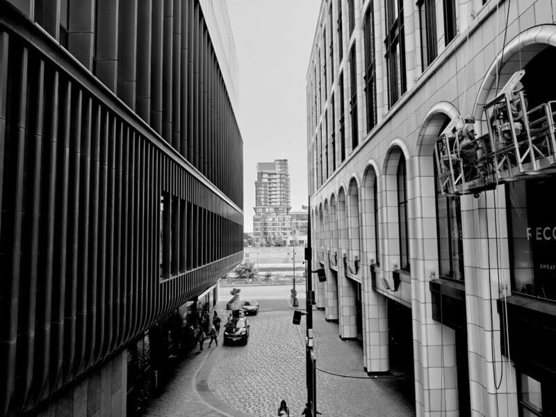 Black and white photo of a city street flanked by modern buildings with arched windows on the right and dark panels on the left, receding toward a distant apartment building. A few people and cars are visible below.