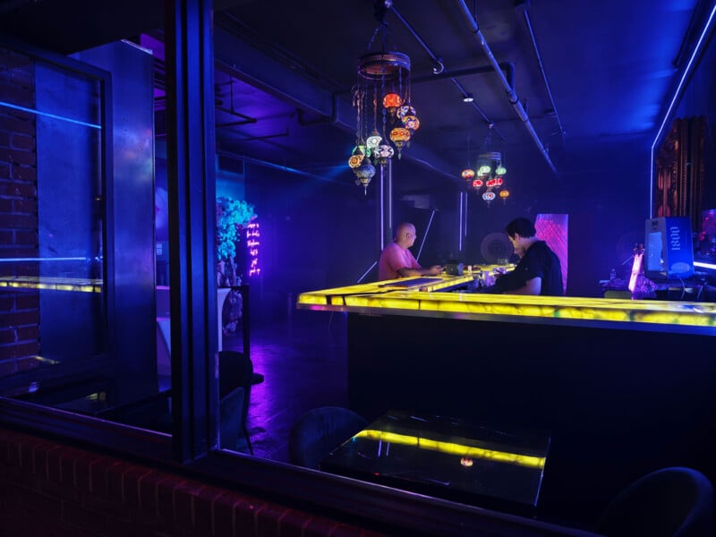 A dimly lit bar with neon lighting features two people sitting at a glowing yellow bar counter, colorful hanging lamps above, and purple-blue ambient lights creating a vibrant, modern nightlife atmosphere.