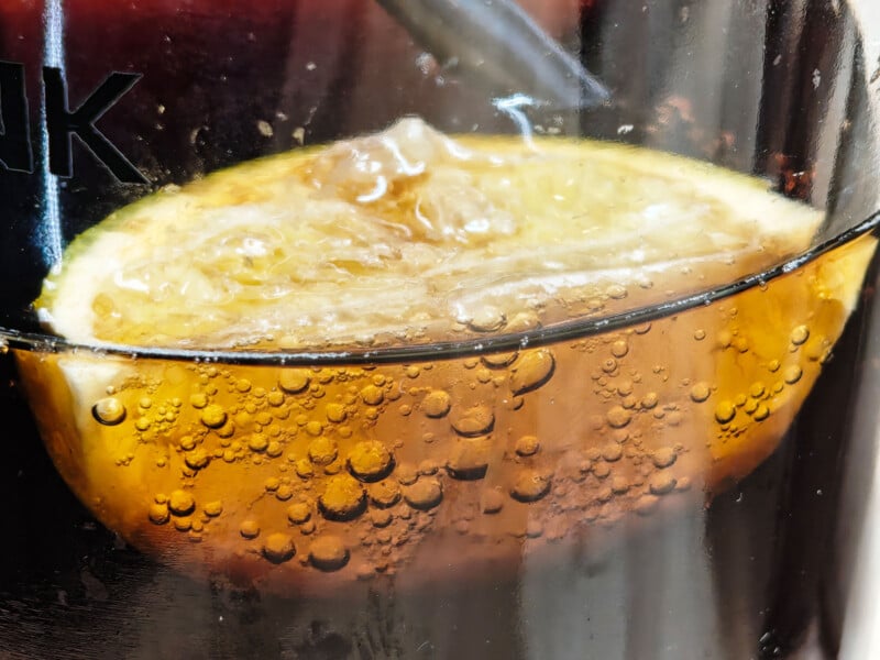 Close-up of a slice of lemon in a fizzy brown drink, with bubbles clinging to the lemon and glass, creating a refreshing and vibrant appearance.