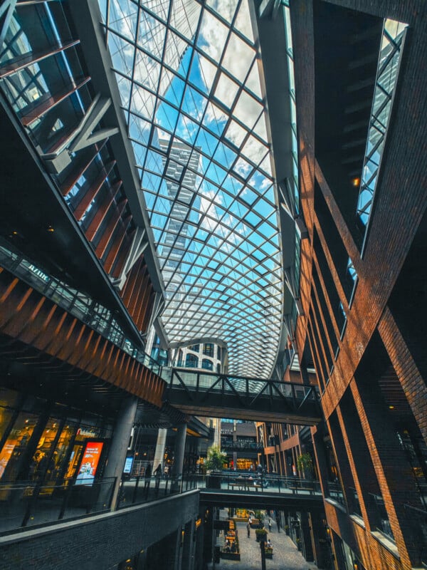 A modern indoor shopping mall with multiple levels, tall brick walls, and a large glass ceiling letting in natural light. The architecture features geometric patterns and walkways connecting different areas.
