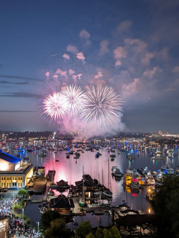 Colorful fireworks explode in the night sky over a marina filled with boats, reflecting on the water. Crowds gather near waterfront buildings to watch the celebration. City lights illuminate the background.