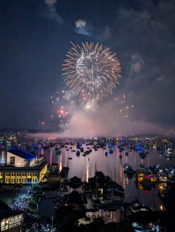 Fireworks burst in the night sky above a marina filled with boats, their lights reflecting on the water. People are gathered near a building at the waterfront, watching the celebration.