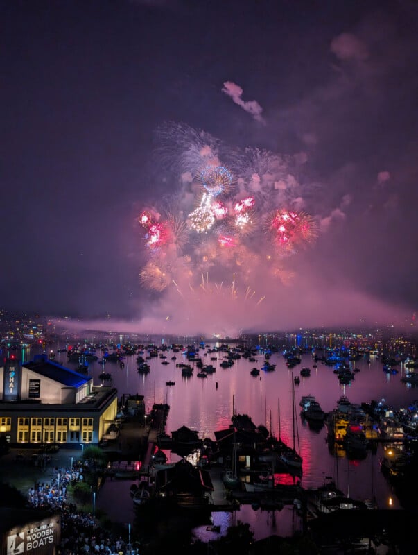 A vibrant display of colorful fireworks lights up the night sky over a harbor filled with boats, their reflections shimmering on the water. City lights and a waterfront building are visible in the foreground.