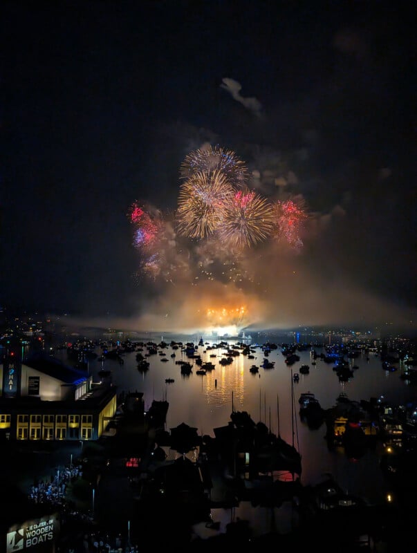 A nighttime scene of a harbor with boats on the water, illuminated by vibrant fireworks in the sky and their reflections. Crowds gather near buildings along the waterfront, watching the colorful display.