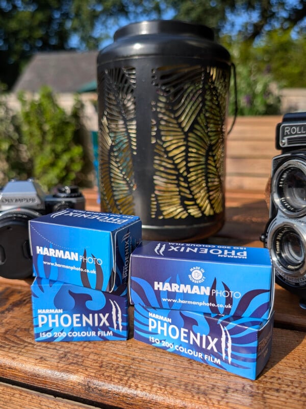 Two boxes of Harman Phoenix color film sit on a wooden table with vintage cameras and a black lantern behind them, outdoors in bright sunlight.