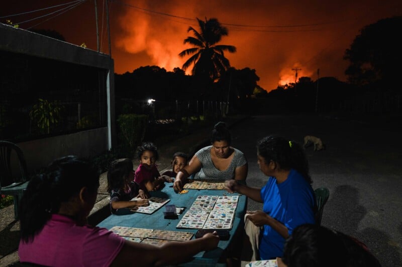 A group of people, including children, play a board game at a table outdoors at night. An orange glow from a wildfire is visible in the background, with palm trees and smoke silhouetted against the sky.