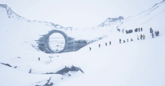 A group of people hike in a line up a snowy slope toward a large circular structure resembling a ring, set amid a vast, snow-covered mountainous landscape.