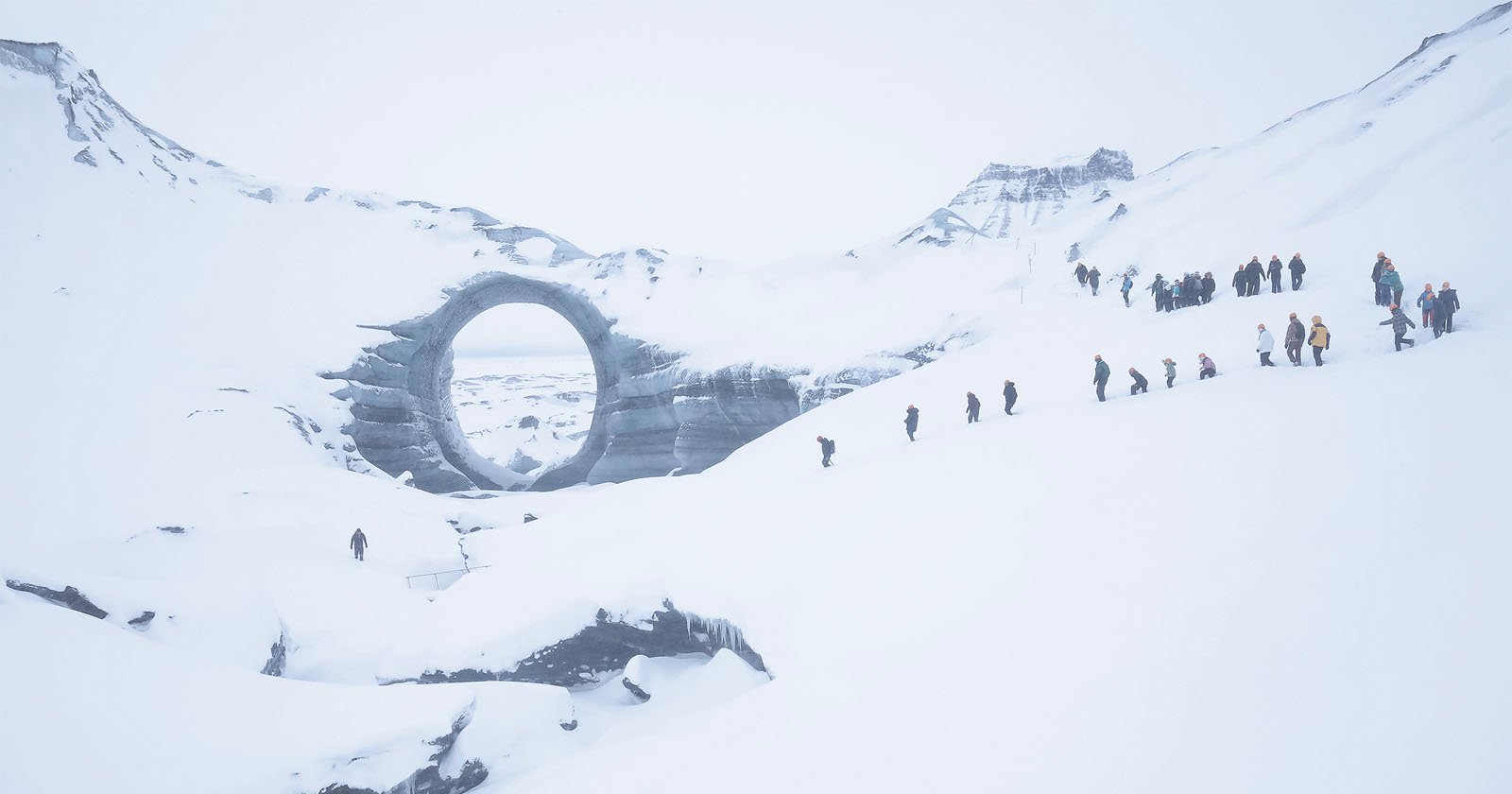 A group of people hike in a line up a snowy slope toward a large circular structure resembling a ring, set amid a vast, snow-covered mountainous landscape.