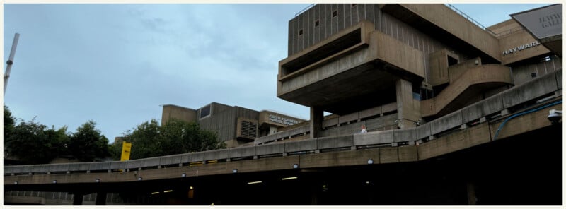 A wide view of the Hayward Gallery in London, featuring a prominent brutalist concrete structure with geometric shapes under a cloudy sky. Trees and signage are visible around the building.