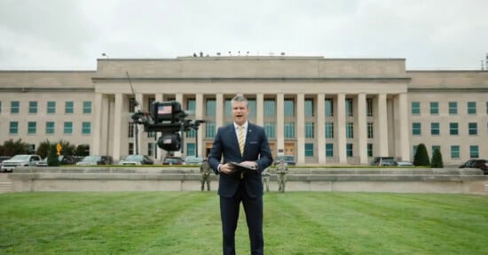 A man in a suit stands on grass in front of a large government building, speaking while holding papers. A black drone hovers in front of him, and several uniformed soldiers are visible in the background.