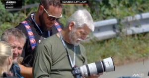 Two men with cameras, one with a large telephoto lens, review photos outdoors near a racetrack during a MotoGP event. A "WARM UP LAP" graphic is displayed at the top of the image.