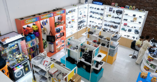 A brightly lit camera shop displays various cameras, lenses, and accessories on shelves. Two people sit at a counter on the right, while colorful bags and gear are organized on the left. The store has white floors and brick walls.