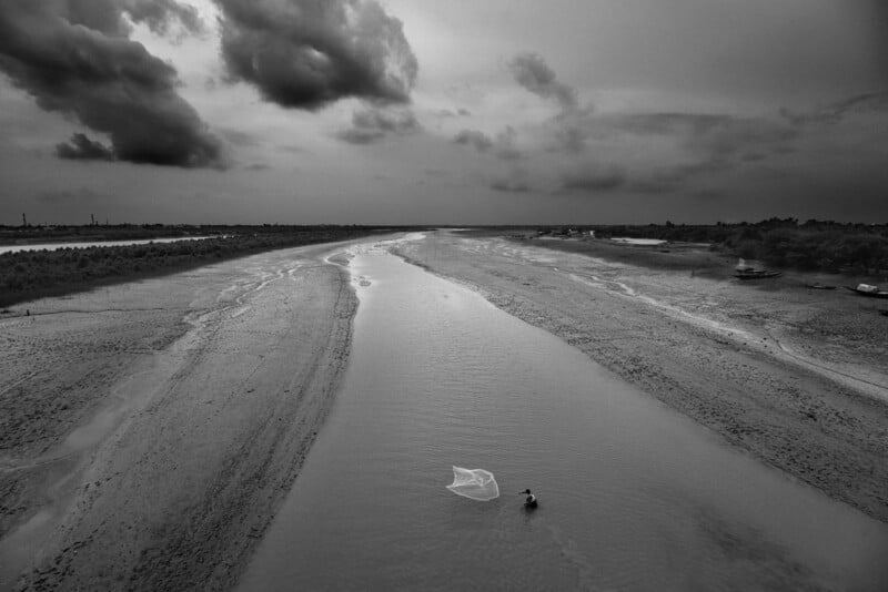 A lone person casts a fishing net in the shallow water of a wide, mostly dry riverbed under a cloudy sky, with stretches of sand and sparse vegetation on both sides. The scene is in black and white.