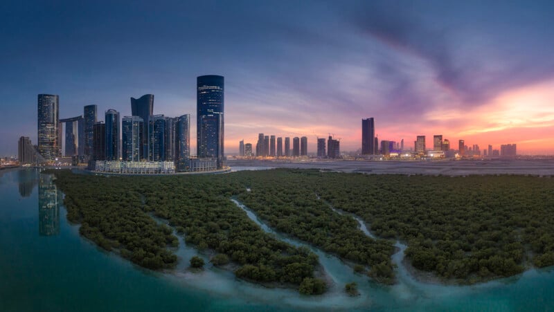 A panoramic view of a modern city skyline at sunset, with tall skyscrapers reflected in calm water and dense green mangroves in the foreground under a colorful sky.
