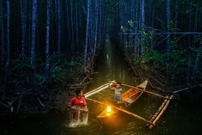 Two people navigate a narrow, tree-lined waterway at dusk; one rows a small wooden boat with a lantern while the other wades beside it, surrounded by dense mangrove trees.