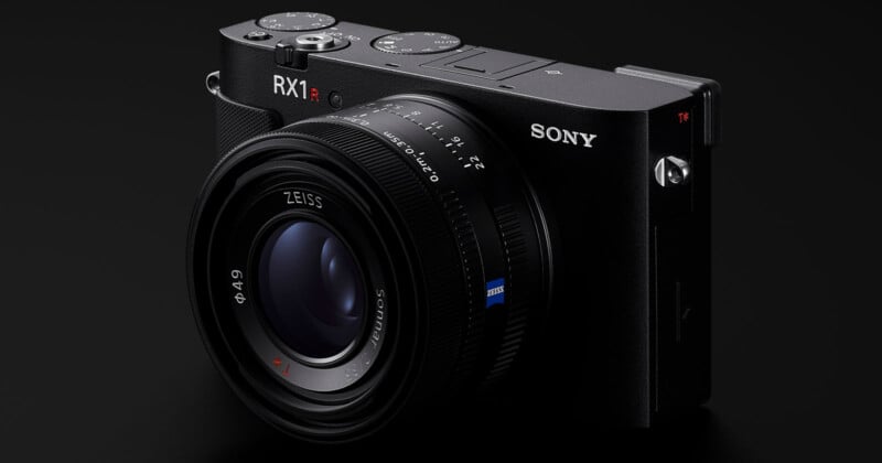 A close-up of a black Sony RX1R II digital camera with a Zeiss lens, showing detailed buttons and dials on a dark background.