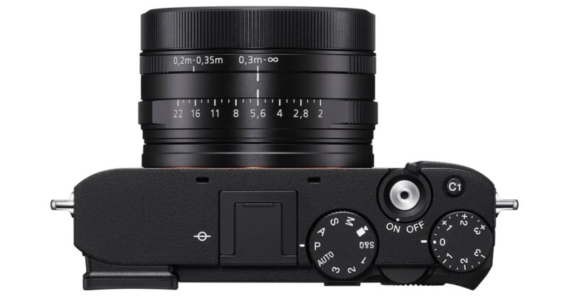 Top view of a black digital camera showing the lens with focus and aperture markings, a mode dial, power switch, shutter button, and other control buttons.