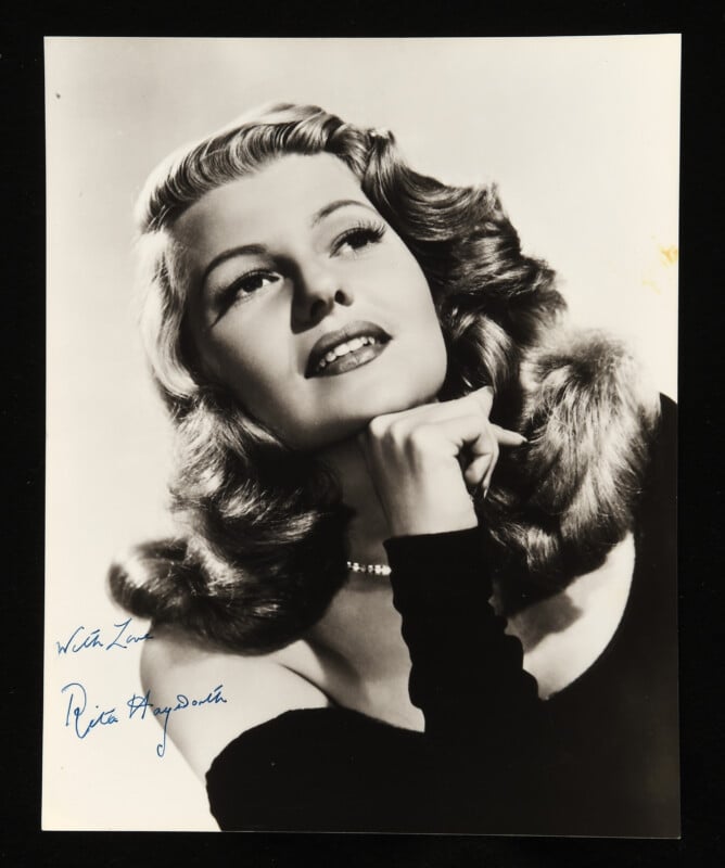 Black-and-white portrait of a glamorous woman with wavy hair, wearing an off-shoulder dress and a pearl necklace, resting her chin on her hand. A handwritten signature is visible in the lower left corner.