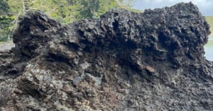 Large, jagged rock formation with rough, dark brown and black surface textures, set outdoors with trees and greenery visible in the blurred background.