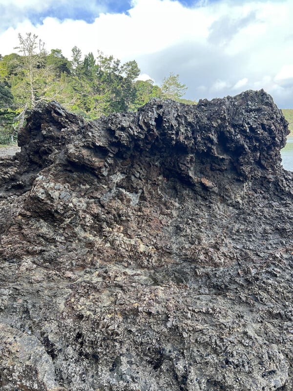 A large, dark, jagged volcanic rock formation sits outdoors, with trees and cloudy sky visible in the background.