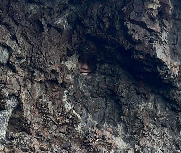 A rocky surface with a small opening reveals part of a person's face, including their eyes and mouth, peeking through a hole in the rock.
