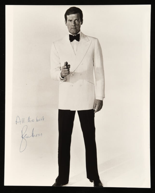 A man in a white tuxedo jacket, black pants, and bow tie stands facing forward, holding a small gun. The photo is signed with the message “All the best” and a signature in blue ink on the left side.