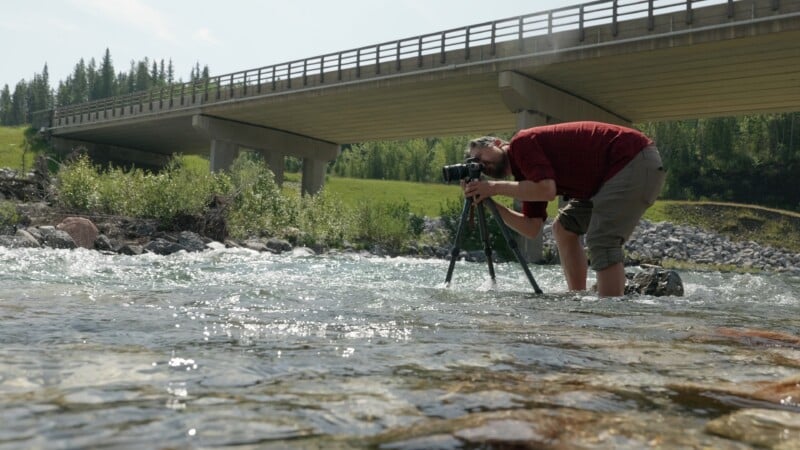 A person wearing a red shirt kneels in a shallow river, using a camera on a tripod. A large concrete bridge spans the river in the background, surrounded by greenery and trees.