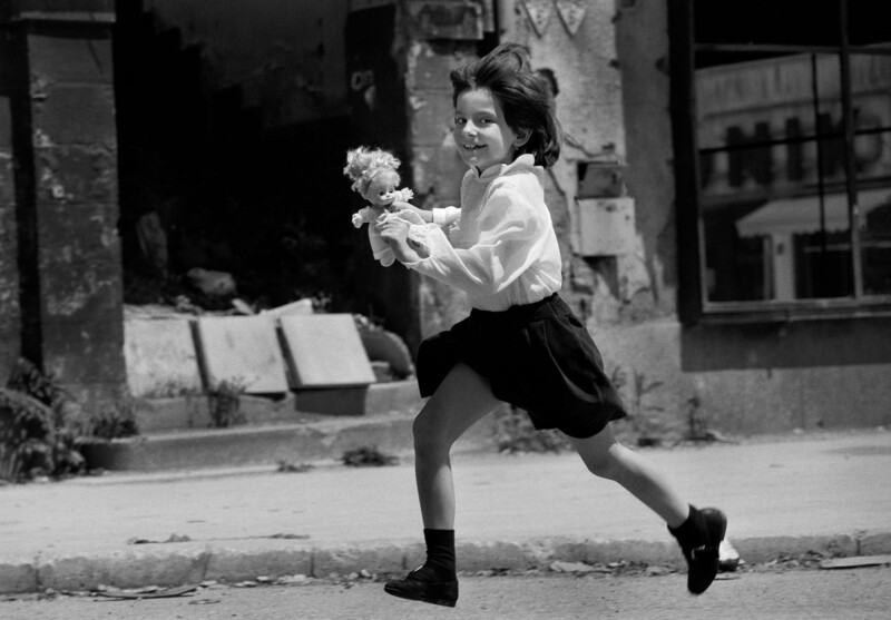A young girl in a skirt and blouse runs joyfully down a street, holding a doll in her hands. The background shows old, damaged buildings and debris, giving a sense of contrast to her playful energy.