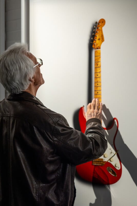 An older man in a leather jacket stands in front of a red electric guitar displayed on a wall, reaching out to touch its neck while looking at it closely.