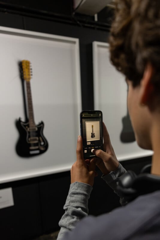 A person takes a photo of a framed black electric guitar on display in a gallery, using a smartphone. The guitar is showcased against a white background.