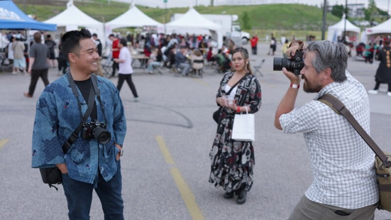 A man in a blue patterned jacket poses for a photo, smiling at the camera, while another man takes his picture. A woman in a long patterned dress and people sitting under tents are visible in the background at an outdoor event.
