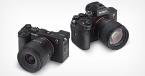 Two Sony Alpha mirrorless cameras with attached lenses are displayed on a white background, one facing forward and the other angled slightly to the side, showing different camera body designs.