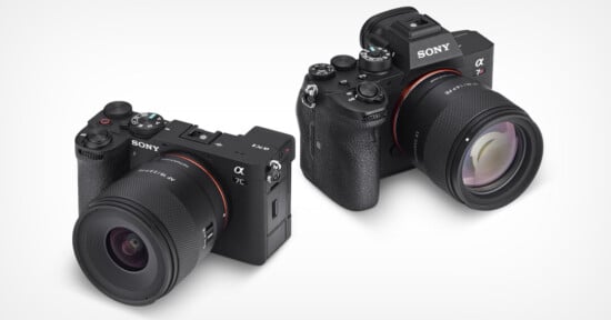 Two Sony Alpha mirrorless cameras with attached lenses are displayed on a white background, one facing forward and the other angled slightly to the side, showing different camera body designs.