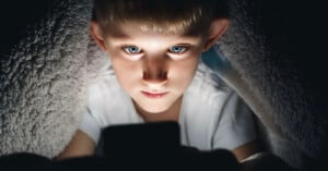 A young boy with blue eyes sits under a blanket, illuminated by the light from a smartphone or tablet he is holding, staring intently at the screen in a dark environment.