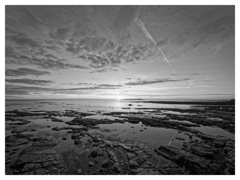 Black and white photo of a rocky shoreline at low tide, with pools of water reflecting the cloudy sky. The sun is low on the horizon, creating a dramatic contrast and a peaceful, expansive seascape.