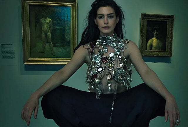 A woman with long dark hair wears an ornate, jewel-covered top and black pants, sitting with arms outstretched in front of two framed paintings of nude figures in a dimly lit art gallery.