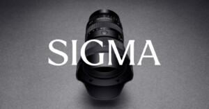 A Sigma camera lens with a lens hood is positioned vertically on a gray surface, with the word "SIGMA" in large white letters overlaid across the image.