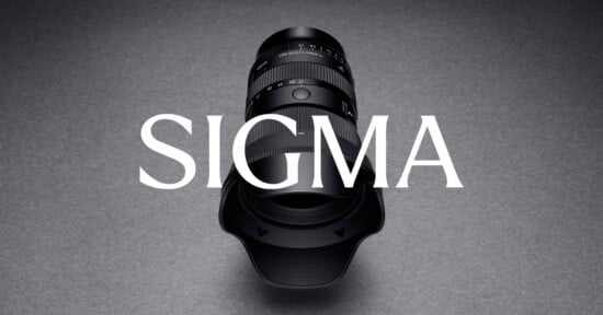 A Sigma camera lens with a lens hood is positioned vertically on a gray surface, with the word "SIGMA" in large white letters overlaid across the image.