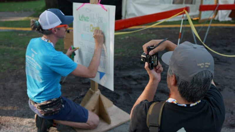A person in a blue shirt kneels while writing on a large board, as another person wearing a hat crouches nearby, taking a photo of the board with a camera. They are outdoors near caution tape and a tent.