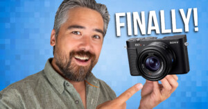 A smiling man with gray hair and a beard points to a Sony RX1 camera with the word "FINALLY!" in bold white letters on a blue pixelated background.